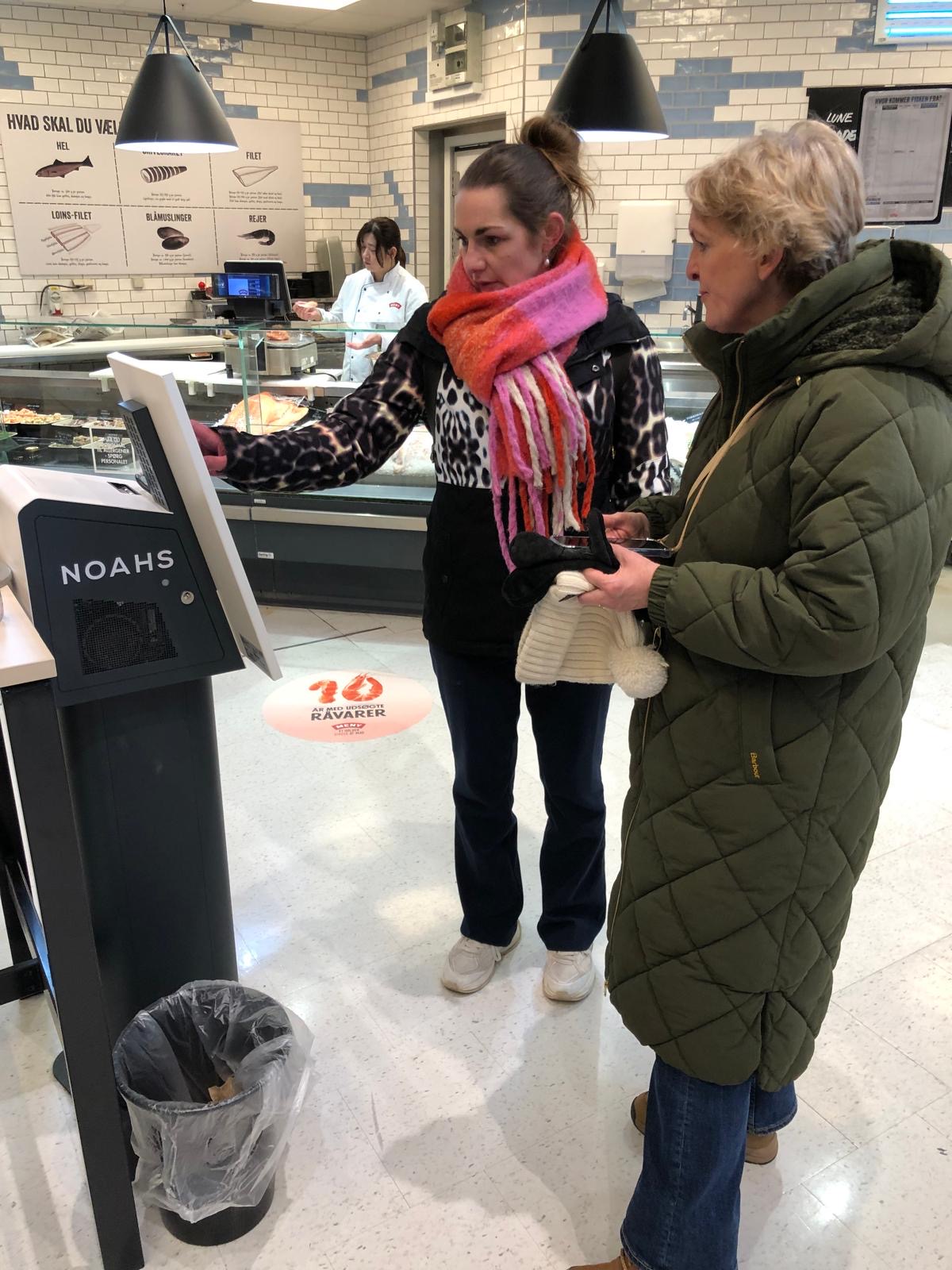 Maaike de Reuver en Jennifer Westgeest bij een bestelzuil met verse maaltijden in een Meny-supermarkt
